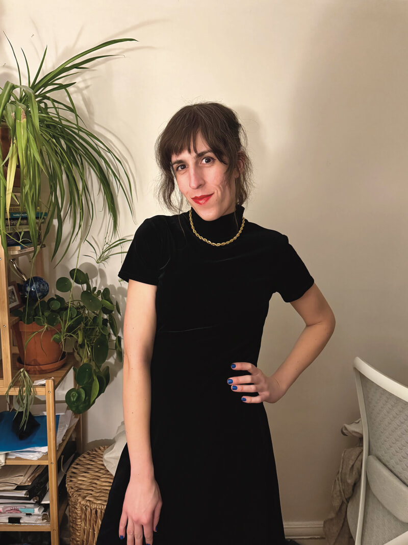 I'm wearing a black velour dress, red lipstick, and a golden necklace. My nails are painted blue, and my left hand is on my hip.