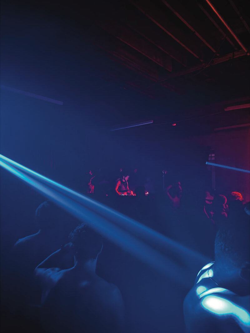 In a dark room iluminated by a cool blue laser, indistinct people stand facing a DJ, lit up by a red light.