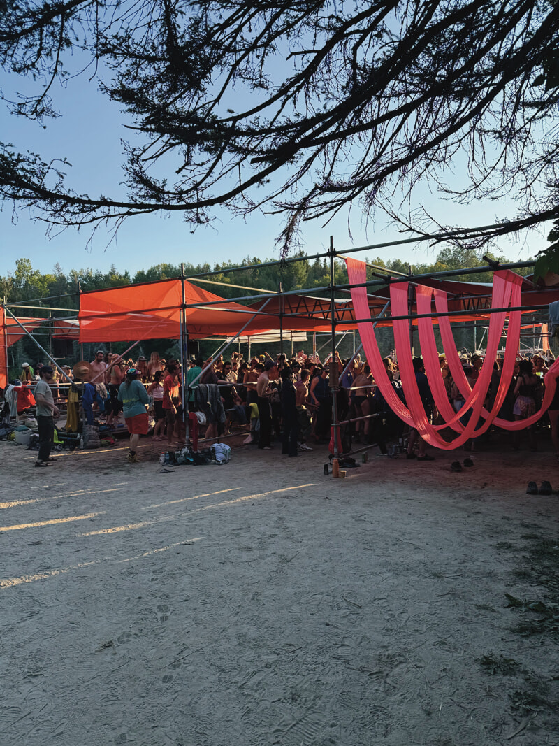 Hundreds of people are dancing on the beach surrounded by a temporary scaffolding structure which large pieces of orange fabric loosely hang.